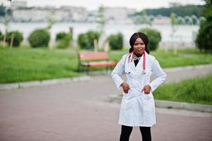 joven doctora afroamericana en bata blanca con un estetoscopio posado al aire libre. foto