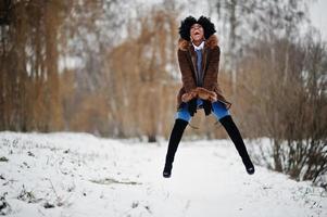 Curly hair african american woman wear on sheepskin coat and gloves posed at winter day, having fun and jump. photo