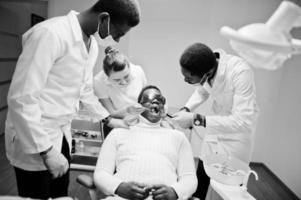 Multiracial dentist doctors team. African american man patient at UV protective glasses. His teeth treated with the help of a dental UV curing light lamp and a dental mirror. photo