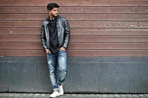 Stylish and casual asian man in black leather jacket, headphones posed against wooden wall. photo