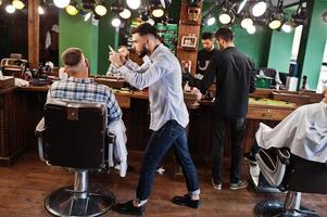 Handsome bearded man at the barbershop, barber at work, making photo on his phone.