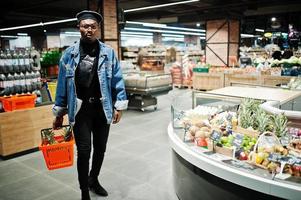 Elegante hombre afroamericano casual con chaqueta de jeans y boina negra sosteniendo una canasta con piñas en la sección orgánica de frutas del supermercado. foto