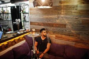 hombre árabe de barba elegante con gafas y camiseta negra fumando narguile en el bar interior. modelo árabe descansando. foto