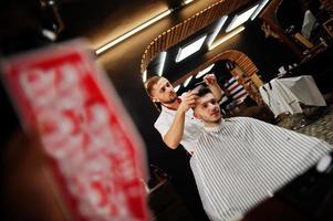 Young bearded man getting haircut by hairdresser while sitting in chair at barbershop. Barber soul. photo