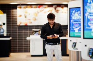 Stylish indian man posed at fast food cafe near self pay floor kiosk with mobile phone at hand. photo