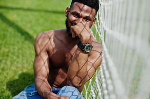 Handsome sexy african american muscular bare torso man at jeans overalls posed at green grass agasinst football gates of stadium field. Fashionable black man portrait. photo