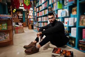 estudiante árabe alto e inteligente, vestido con chaqueta negra de jeans y anteojos, en la biblioteca sentado contra estantes de libros. foto