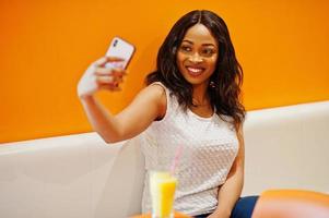 African woman sitting on cafe against orange wall with pineapple juice and mobile phone in hands, she making selfie. photo