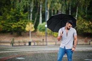 hombre de barba árabe alto de moda usa camisa, jeans y gafas de sol con paraguas posado bajo la lluvia en la plaza del parque. foto