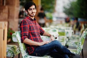 Young indian student man at red checkered shirt and jeans sitting at evening cafe. photo