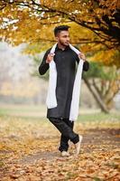 hombre indio con estilo en ropa tradicional negra con bufanda blanca posada al aire libre contra el árbol de hojas amarillas de otoño. foto