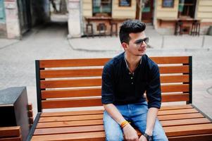 elegante modelo indio con ropa informal, camisa negra y gafas de sol posado al aire libre en la calle de la india y sentado en un banco. foto