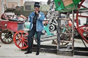 hombre afroamericano con chaqueta de jeans, boina y anteojos, fumando cigarro y posó contra un viejo vehículo retro y mirando el teléfono. foto