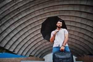 el hombre de barba árabe alto de moda usa camisa, jeans y gafas de sol con paraguas y bolso de caja posado en el salón de la arena. foto