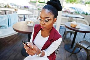 African american girl wear in glasses with mobile phone sitting at outdoor caffe. photo