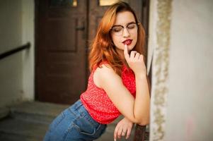 Attractive redhaired woman in eyeglasses, wear on red blouse and jeans skirt posing. photo