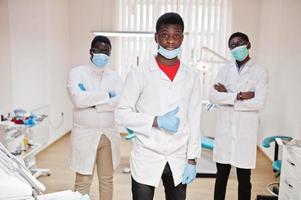 Three african american male doctor with crossed arms in dental clinic. Show thumb up. photo