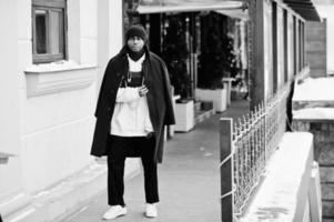 Stylish urban style african american man in black coat and hat, pink hoodie posed at winter day. photo