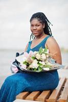 African american dark skinned plus size model posed in a blue shiny dress with bouquet of flowers sitting on bench against sea side. photo