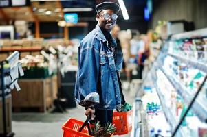 elegante hombre afroamericano casual con chaqueta de jeans y boina negra sosteniendo dos canastas, parado cerca de la nevera y comprando en el supermercado. foto