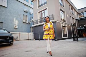 Stylish african american women in yellow jacket posed and earphones on street against modern building. photo