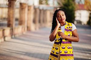 Stylish african american women in yellow jacket posed on street with hot drink in disposable paper cup. photo