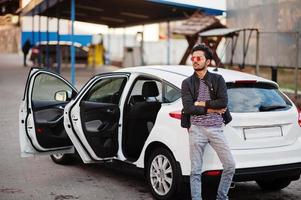 South asian man or indian male wear red eyeglasses stand near his white transportation on car wash. photo