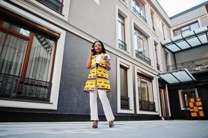 Stylish african american women in yellow jacket posed on street with hot drink in disposable paper cup and mobile phone at hands. photo