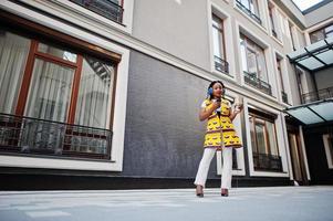 Stylish african american women in yellow jacket posed and earphones on street against modern building with hot drink in disposable paper cup. photo