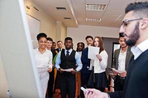 Arabian business coach presenting report standing near whiteboard pointing on sales statistic shown on diagram and chart teach diverse multiracial company members gathered together in conference room. photo