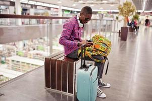 hombre afroamericano con camisa a cuadros, gafas de sol y jeans con maleta y mochila. el viajero negro revisó su pasaporte en la tienda libre de impuestos. foto