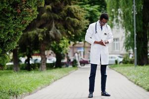 Stylish african american doctor with stethoscope and lab coat, at glasses posed outdoor. photo