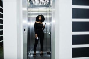moda joven y hermosa mujer de negocios afroamericana con peinado afro en elegante negro, de pie en el ascensor con el teléfono y la computadora portátil en las manos. foto