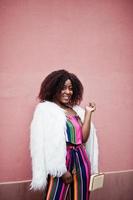 Fashionable african american woman in striped jumpsuit with fluffy faux fur coat and handbag posed against pink wall. photo