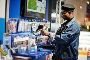 elegante hombre afroamericano casual con chaqueta de jeans y boina negra usando gafas vr en la tienda de electrónica. foto