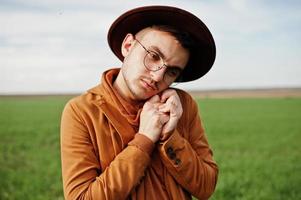 Stylish man in glasses, brown jacket and hat posed on green field. photo