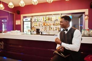 joven y apuesto hombre africano con camisa blanca, chaleco negro y corbata de moño con monedero posado contra el mostrador del bar en el club nocturno. foto