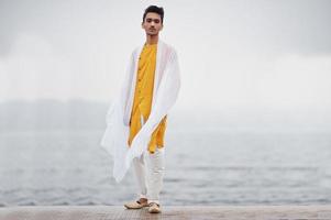 hombre indio con estilo en ropa tradicional amarilla con bufanda blanca posada al aire libre contra el muelle de niebla del mar. foto