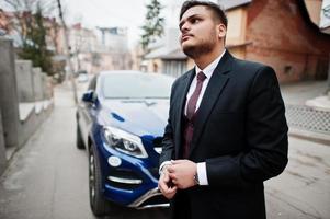 Rich indian businessman in formal wear standing against business suv car. photo