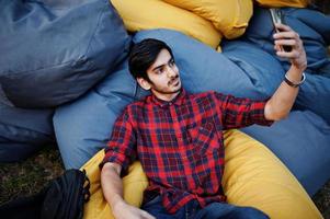 joven estudiante indio con camisa a cuadros y jeans sentado y relajado en almohadas al aire libre y haciendo selfie. foto