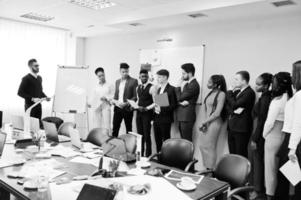 Mixed race business coach presenting report standing near whiteboard pointing on sales statistic shown on diagram and chart teach diverse company members gathered together in conference room. photo