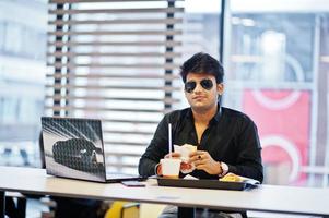 Stylish indian man in sunglasses sitting at fast food cafe against his laptop. photo