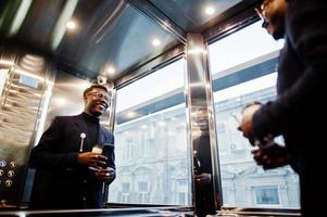 Fashionable african american man in suit and glasses with mobile phone and cup of coffee at hands posed inside elevator. photo
