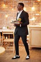 Respectable young african american man in black suit hold tray with double burger against brick wall of restaurant with lights. photo