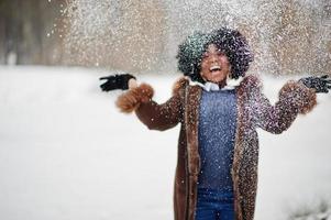 Curly hair african american woman wear on sheepskin coat and gloves posed at winter day throws up snow. photo