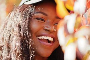 Stylish african american girl in cap posed at sunny autumn day against red leaves. Africa model woman. photo