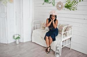 bonita mujer afroamericana con anteojos y sombrero posada en la habitación sentada en un sofá vintage. foto