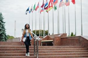 African student female posed with backpack and school items on yard of university, against flags of different countries. photo