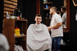 un joven barbudo que se corta el pelo con un peluquero mientras se sienta en una silla en la barbería. alma de barbero. foto