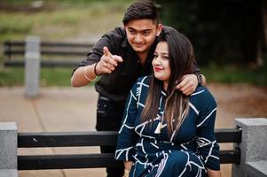 Love story of indian couple posed outdoor, sitting on bench together. Show finger photo